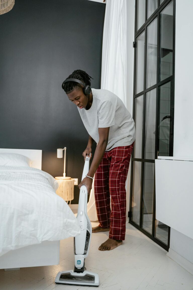 A young man cleans a bedroom with a vacuum cleaner, wearing headphones and pajamas.