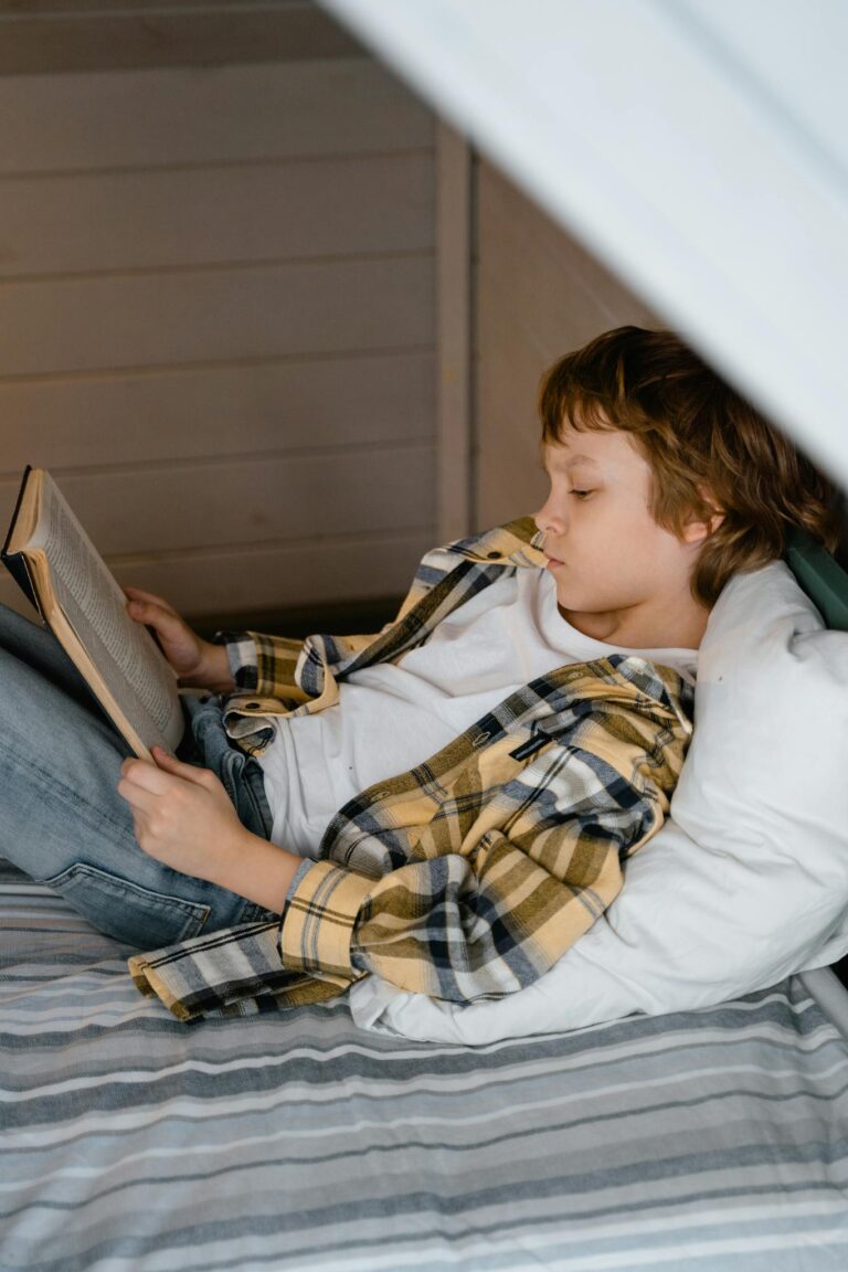 Young boy in casual clothing reading a book indoors, resting on a bed.